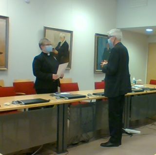 Salla Häkkinen vastaanottaa onnitteluja piispa Matti Revolta suoritettuaan pastoraalitutkinnon. Hei seisovat vastakkaisilla puolilla pöytää maskit kasvojensa suojana.