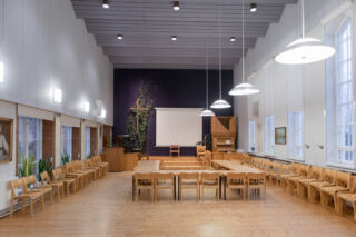 Kangasalan kirkonkylän seurakuntakodin valoisa ja monikäyttöinen seurakuntasali jossa vaaleat ikkunaseintä, puunvärinen lattia, seiniä kiertämässä puisten tuolien rivistöt ja keskellä pöytä- ja tuoliryhmät.