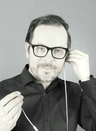 Jukka Saksi mustasankaisissa silmälaseissa pitelee käsissään kuulokkeita, toinen kuuloke korvalle kohotettuna.
