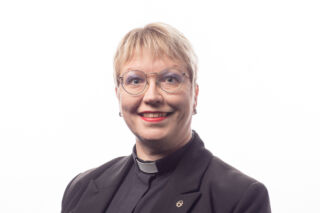 Salla Häkkinen valkoista taustaa vasten yllään papin puku ja tunnukset.