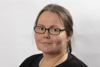 Maarit Vuolabilla on ruskeat hiukset ja tummasankaiset silmälasit, yllään hänellä on musta paita. Hän katsoo kameraan hento hymy huulillaan.