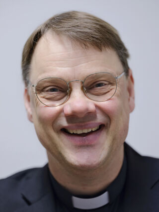 Miikka Anttila kasvoillaan naurava ilme, mustassa paidassa papin valkoinen "sokeripala" kaulassa. Anttilalla on ruskeat lyhyet hiukset ja silmälasit. Tausta on vaalea.