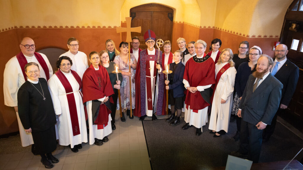 Piispa Matti Repo piispankaavussa ja piispansauva kädessä ympärillään diakonian virkaan ja pappisvirkaan vihityt, muut UTV-valmennukseen osallistuneet ja messun avustajat. Taustalla keltasävyinen seinä ja ruskea ovi.