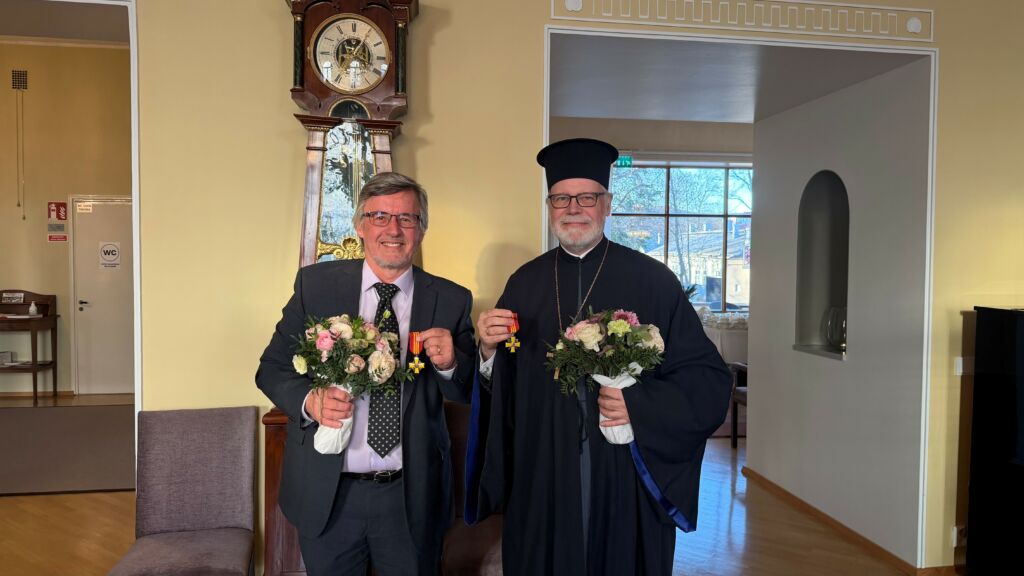 Pekka Simojki ja Heikki Huttunen seisovat hymyillen suuren kaappikellon edessä, takana avautuu ovi suureen ja valoisaan saliin. Kädessään he molemmat pitelevät kunniamerkkejään.