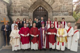 Ryhmä pappeja ja seurakuntalaisia poseeraa kivisen kirkon sisäänkäynnin edessä. Eturivissä on pappeja punaisissa, valkoisissa ja kultaisissa liturgisissa asuissa; keskellä seisoo piispa Matti Repo hiippa päässään ja sauva kädessään. Taustalla seisoo joukko ihmisiä papinpuvuissa ja juhlavaatteissa.